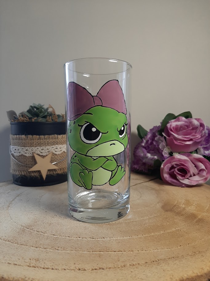 Verre haut transparent avec une petite grenouille verte rigolote qui boude, portant un grand nœud rose sur la tête, peinture sur verre artisanale Aleco.
