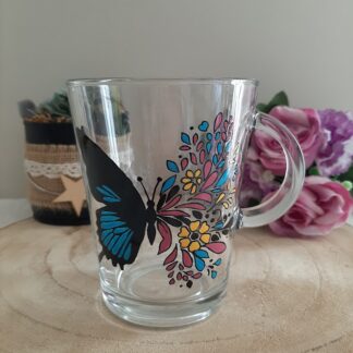 Mug en verre transparent décoré d'un papillon dont l'aile droite se transforme en fleurs bleues, jaunes et roses, peint à la main, artisanat Aleco Vendée.