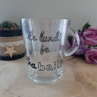 Mug en verre transparent avec anse, portant l'inscription humoristique peinte à la main en noir : "le lundi je tra-baille", avec le mot "baille" mis en évidence, artisanat Aleco.