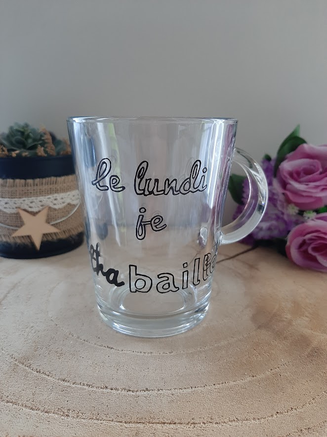 Mug en verre transparent avec anse, portant l'inscription humoristique peinte à la main en noir : "le lundi je tra-baille", avec le mot "baille" mis en évidence, artisanat Aleco.