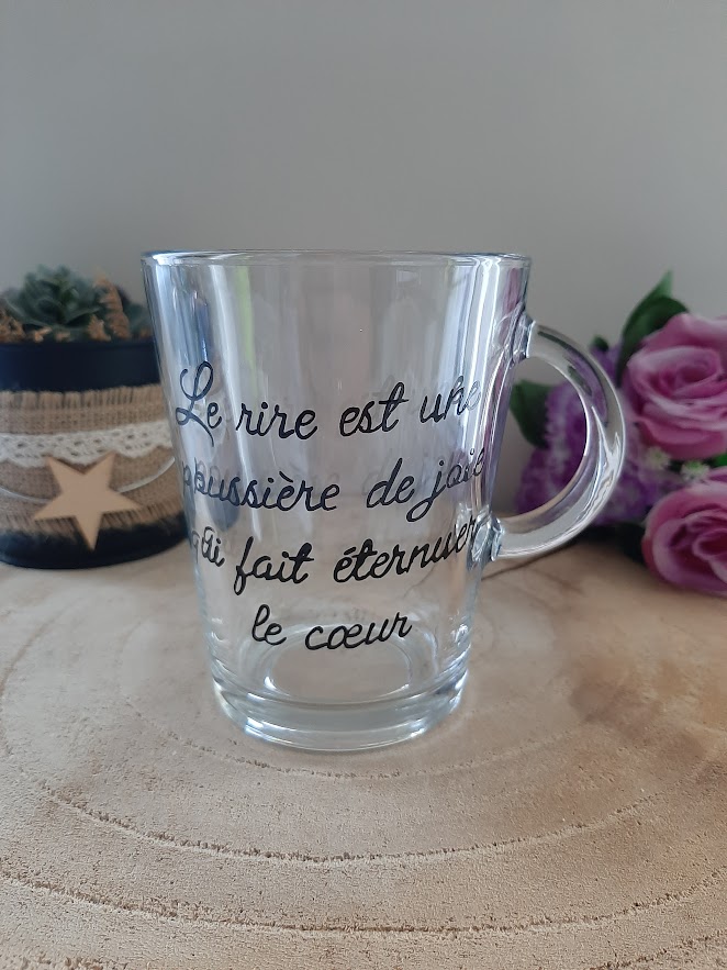 Mug en verre transparent avec anse, portant la citation poétique calligraphiée à la main en noir : "Le rire est une poussière de joie qui fait éternuer le cœur", artisanat Aleco.