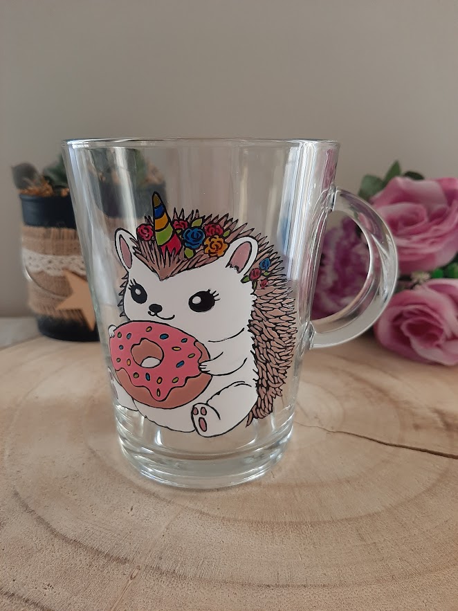 Mug en verre transparent avec anse, décoré d'une illustration artisanale colorée représentant un hérisson blanc avec une corne de licorne arc-en-ciel, une couronne de fleurs et tenant un donut rose, peint à la main par Aleco.