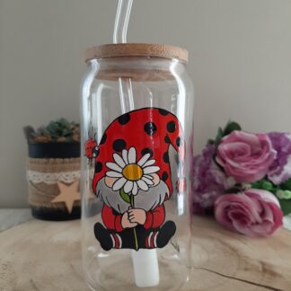 Gobelet en verre transparent avec couvercle en bambou et paille en verre, décoré d'une illustration peinte à la main d'un gnome assis portant un grand bonnet rouge à pois noirs, tenant une grande marguerite blanche, avec une petite coccinelle sur le côté, artisanat Aleco.