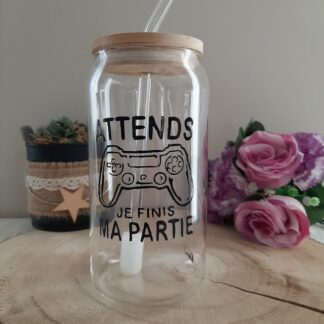 Gobelet en verre transparent avec couvercle en bambou et paille en verre, décoré d'une illustration noire peinte à la main représentant une manette de console de jeux vidéo et l'inscription "ATTENDS JE FINIS MA PARTIE", artisanat Aleco.