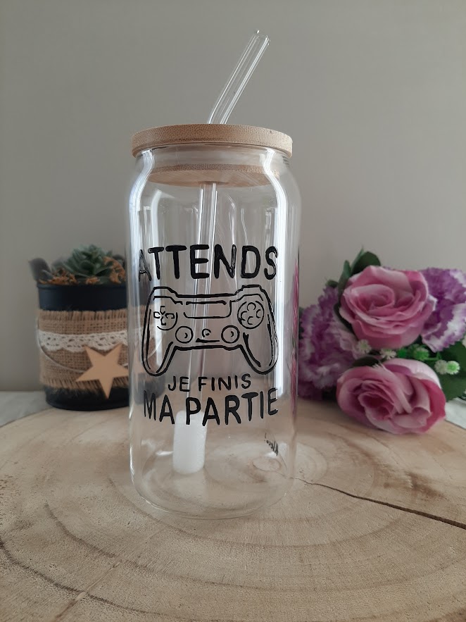 Gobelet en verre transparent avec couvercle en bambou et paille en verre, décoré d'une illustration noire peinte à la main représentant une manette de console de jeux vidéo et l'inscription "ATTENDS JE FINIS MA PARTIE", artisanat Aleco.