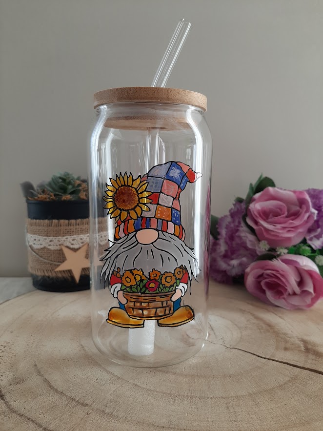 Gobelet en verre transparent avec couvercle en bambou et paille en verre, décoré d'une illustration peinte à la main d'un gnome barbu tenant un panier de fleurs oranges et portant un grand bonnet multicolore style patchwork orné d'un tournesol, artisanat Aleco.