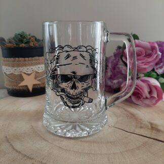 chope à bière en verre transparent avec anse, décorée d'une illustration noire peinte à la main représentant un crâne portant un chapeau bob (bucket hat), fumant un cigare avec un large nuage de fumée en arrière-plan, artisanat Aleco.