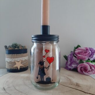 Pot en verre transparent contenant des bougies beiges, doté d'un couvercle noir avec un bougeoir intégré, décoré d'une silhouette peinte à la main d'une petite fille avec une valise tenant trois ballons rouges en forme de cœur, artisanat Aleco.