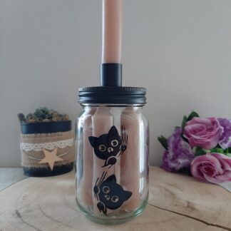 Pot en verre transparent contenant des bougies beiges, doté d'un couvercle noir avec bougeoir intégré, décoré d'une illustration peinte à la main représentant deux petits chats noirs rigolos aux grands yeux expressifs, artisanat Aleco.