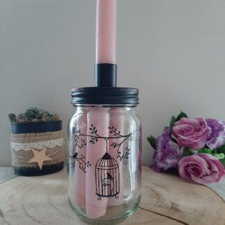 Pot en verre transparent rempli de bougies roses, avec un couvercle noir servant de bougeoir, décoré d'une illustration noire peinte à la main représentant une cage à oiseaux ouverte suspendue à une branche fleurie avec de petits oiseaux, artisanat Aleco.