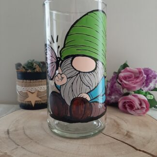 Vase cylindrique en verre artisanal Aleco avec un gnome au bonnet vert assis et un papillon rose posé sur son doigt, peint à la main.