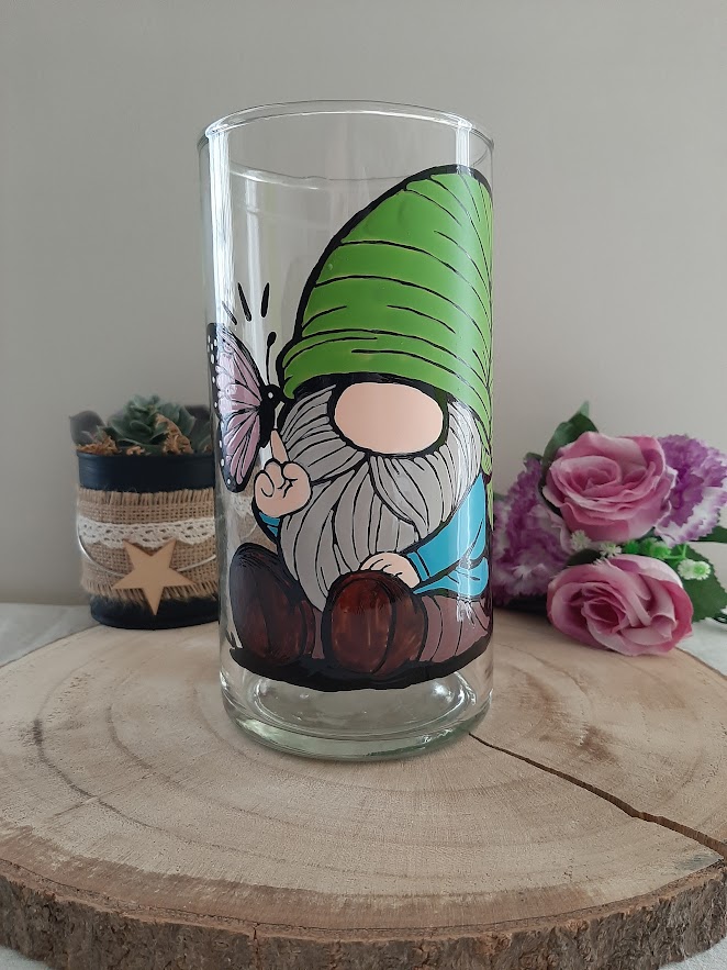Vase cylindrique en verre artisanal Aleco avec un gnome au bonnet vert assis et un papillon rose posé sur son doigt, peint à la main.