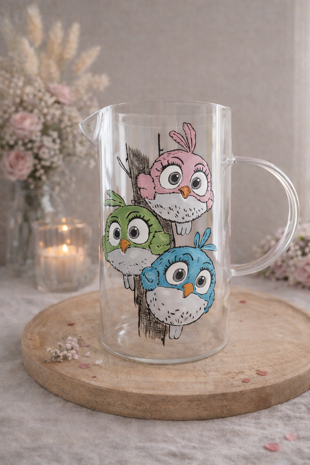 Carafe pichet en verre transparent avec anse, peinte à la main levée avec un motif de trois chouettes et hiboux colorés (rose, vert, bleu) perchés sur un tronc, présentée sur un support en bois