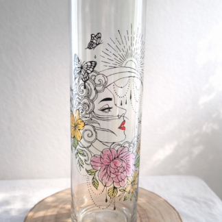 Grand vase cylindrique transparent peint à la main levée avec un motif de profil de femme mystique, des fleurs roses et jaunes, des papillons et une lune, présenté sur un support en bois