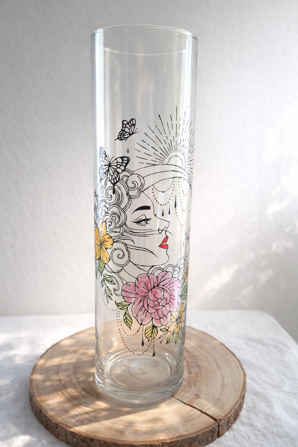 Grand vase cylindrique transparent peint à la main levée avec un motif de profil de femme mystique, des fleurs roses et jaunes, des papillons et une lune, présenté sur un support en bois