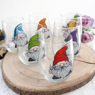 Série de verres gobelets arrondis transparents peints à la main avec des gnomes rigolos aux bonnets colorés (rouge, vert, bleu, orange, jaune, violet), posés sur une tranche de bois