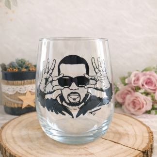 Verre gobelet arrondi transparent peint à la main levée avec une illustration noire représentant le portrait du chanteur Gims avec ses lunettes de soleil, posé sur un support en bois