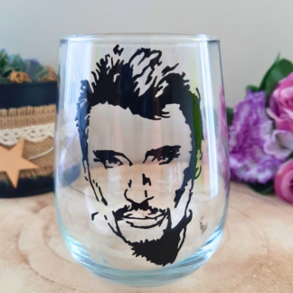 Verre gobelet arrondi transparent peint à la main levée avec une illustration noire représentant le portrait du chanteur Johnny Hallyday, posé sur un support en bois