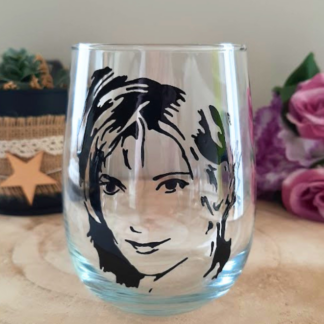Verre gobelet arrondi transparent peint à la main levée avec une illustration noire représentant le visage et le portrait de la chanteuse Mylène Farmer, présenté sur un support en bois