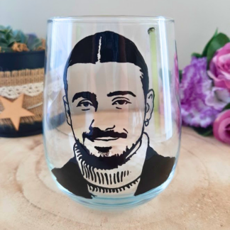 Verre gobelet arrondi transparent peint à la main levée avec une illustration noire représentant le portrait du chanteur Pierre Garnier en col roulé, présenté sur un support en bois