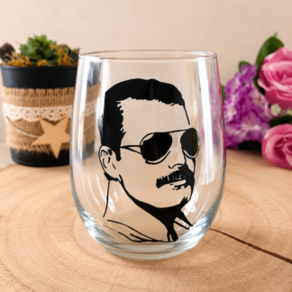 Verre gobelet arrondi transparent peint à la main levée avec une illustration noire représentant le portrait de Freddie Mercury avec ses lunettes de soleil et sa moustache, présenté sur un support en bois