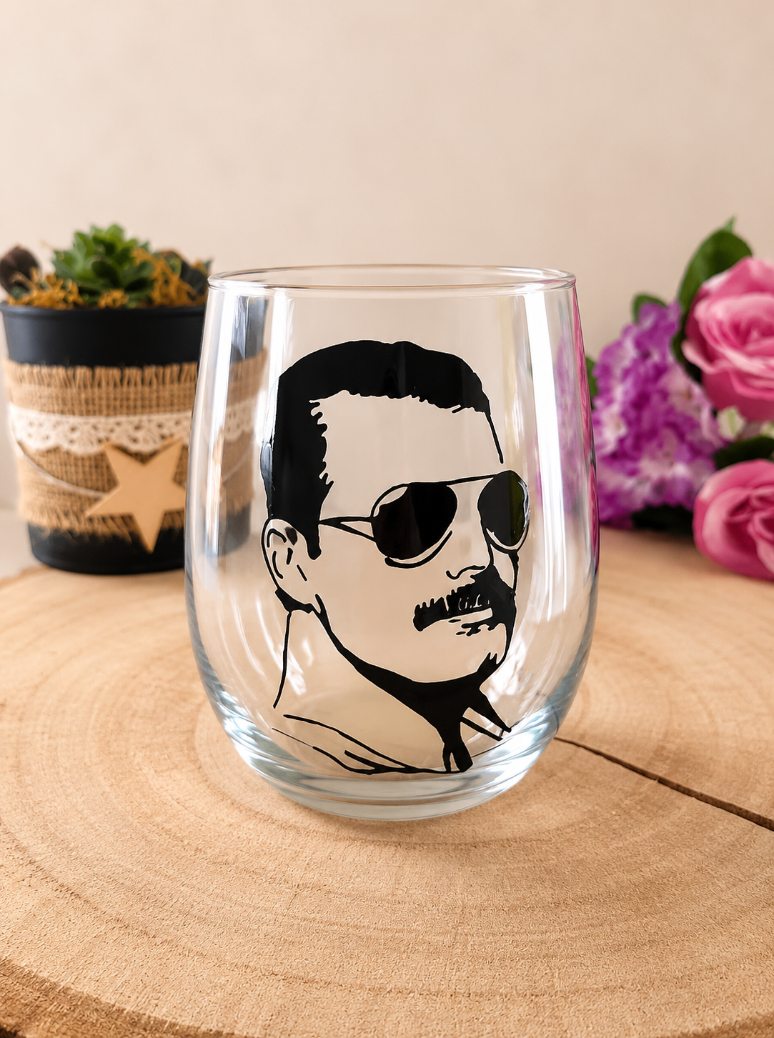 Verre gobelet arrondi transparent peint à la main levée avec une illustration noire représentant le portrait de Freddie Mercury avec ses lunettes de soleil et sa moustache, présenté sur un support en bois