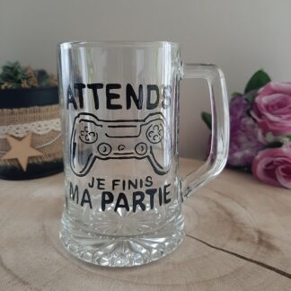 Chope à bière transparente avec une manette de jeu vidéo et le texte "Attends je finis ma partie" peint à la main en noir.