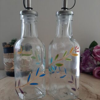 Duo de flacons huilier et vinaigrier en verre transparent peints à la main avec un motif de feuillage bleu, violet et jaune, becs verseurs en inox, posés sur un socle en bois.