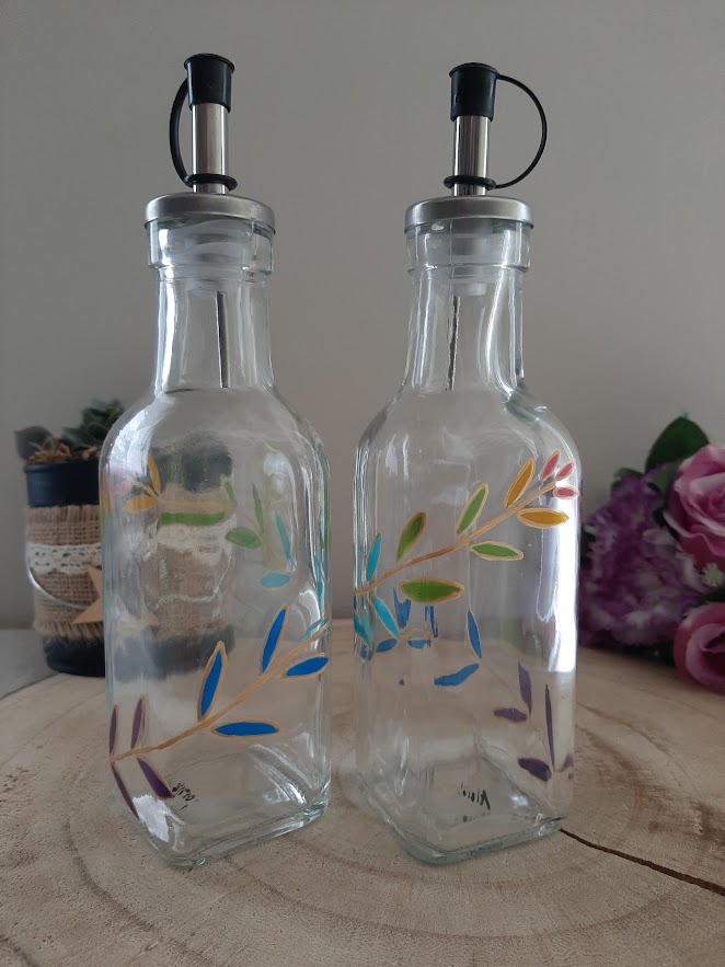 Duo de flacons huilier et vinaigrier en verre transparent peints à la main avec un motif de feuillage bleu, violet et jaune, becs verseurs en inox, posés sur un socle en bois.