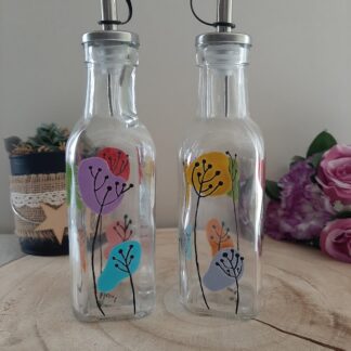 Duo de bouteilles huilier et vinaigrier en verre avec des motifs de fleurs stylisées violettes, jaunes, oranges et turquoises peintes à la main, becs verseurs noirs.