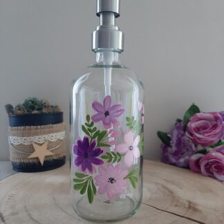 Distributeur de savon en verre transparent avec pompe argentée et décor de fleurs peintes à la main dans les tons roses, violets et mauves.