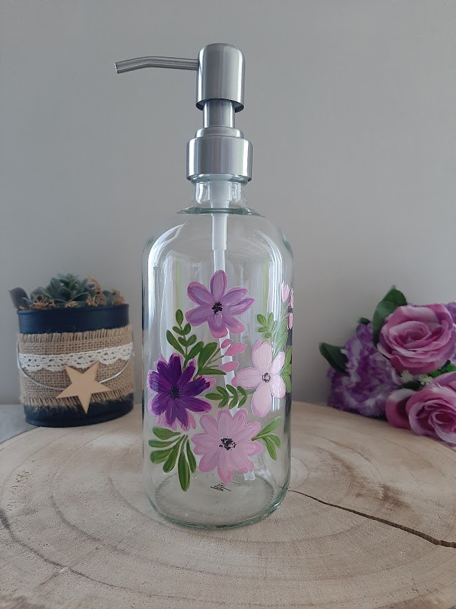 Distributeur de savon en verre transparent avec pompe argentée et décor de fleurs peintes à la main dans les tons roses, violets et mauves.