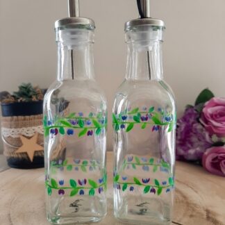 Duo de bouteilles carrées en verre pour huile et vinaigre, peintes à la main avec une frise double de feuilles vertes et petites fleurs bleues et violettes, posées sur un socle en bois