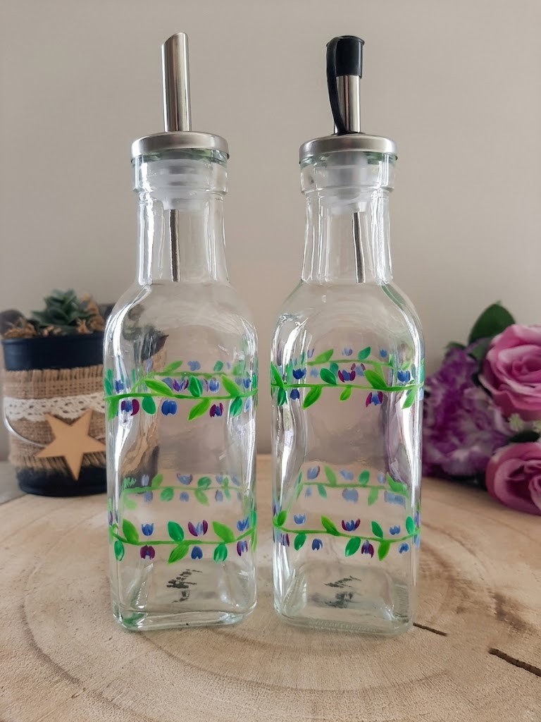 Duo de bouteilles carrées en verre pour huile et vinaigre, peintes à la main avec une frise double de feuilles vertes et petites fleurs bleues et violettes, posées sur un socle en bois