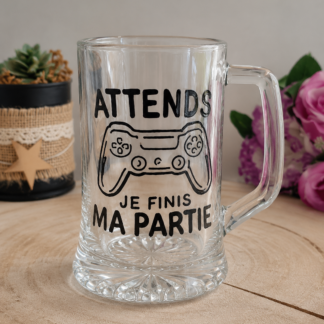 Chope à bière en verre transparent avec anse, peinte à la main avec une manette de jeu vidéo et l'inscription noire Attends je finis ma partie, posée sur un support en bois