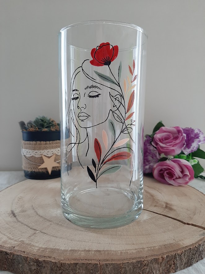 Vase cylindrique en verre de 20 cm de haut, peint à la main avec une illustration en Line Art d'un visage de femme et un coquelicot rouge, posé sur un socle en bois