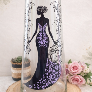Grand vase évasé en verre de 40 cm de haut, peint à la main avec une silhouette de femme de dos en longue robe violette et noire à motifs floraux, posé sur une tranche de bois