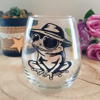 Verre gobelet arrondi transparent peint à la main levée avec une illustration noire de grenouille rigolote portant un chapeau, posé sur un support en bois