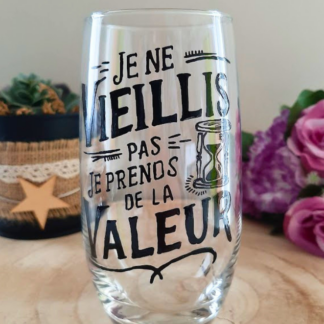 Verre haut transparent peint à la main levée avec une citation humoristique noire sur le temps qui passe et un dessin de sablier, posé sur une tranche de bois