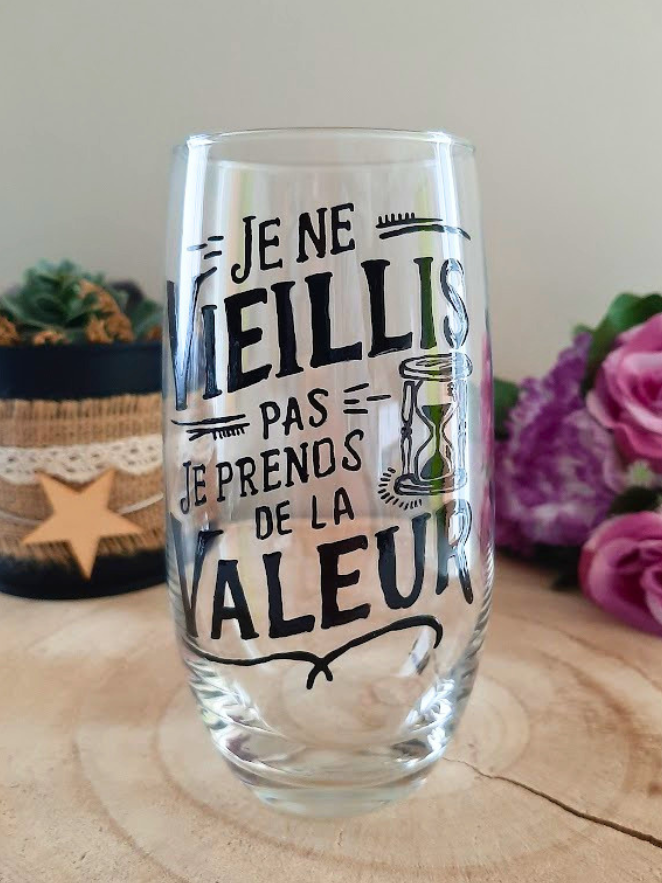Verre haut transparent peint à la main levée avec une citation humoristique noire sur le temps qui passe et un dessin de sablier, posé sur une tranche de bois