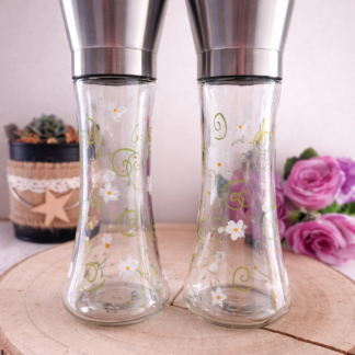Set de deux moulins à sel et poivre en verre peints à la main avec des fleurs blanches et des volutes vertes, posés sur une rondelle de bois