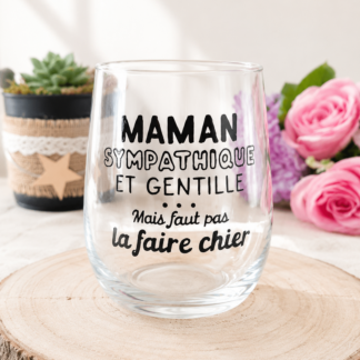 Verre à message humoristique peint à la main « Maman Sympathique et Gentille Mais faut pas la faire chier » posé sur une rondelle de bois avec des roses roses en arrière plan