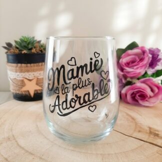 Verre à message peint à la main « Mamie la plus Adorable » avec petits cœurs décoratifs, posé sur une rondelle de bois avec des roses roses en arrière plan