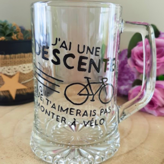 Chope à bière peinte à la main avec jeu de mots humoristique sur le vélo et illustration bicyclette, posée sur une table en bois avec des roses violettes