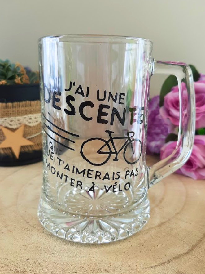 Chope à bière peinte à la main avec jeu de mots humoristique sur le vélo et illustration bicyclette, posée sur une table en bois avec des roses violettes