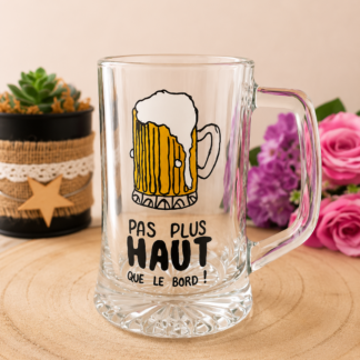 Chope à bière peinte à la main avec illustration chope mousseuse et message humoristique Pas Plus Haut que le Bord, posée sur une table en bois avec des roses violettes