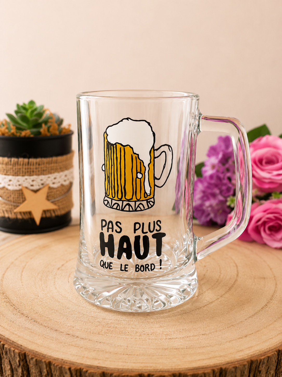 Chope à bière peinte à la main avec illustration chope mousseuse et message humoristique Pas Plus Haut que le Bord, posée sur une table en bois avec des roses violettes