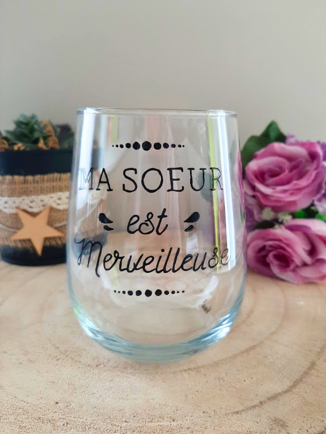 Gobelet en verre peint à la main avec inscription calligraphiée "Ma sœur est merveilleuse" et petits oiseaux noirs, posé sur une table en bois avec des roses roses