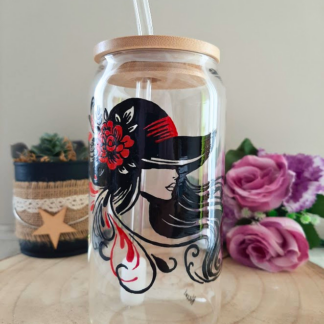 Gobelet paille en verre peint à la main avec silhouette de femme élégante au chapeau et rose rouge, couvercle en bambou et paille en verre, posé sur une table en bois avec des roses roses