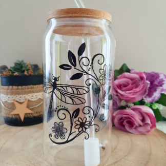 Gobelet paille en verre peint à la main avec libellule et motifs floraux, couvercle en bambou et paille en verre, posé sur une table en bois avec des roses roses